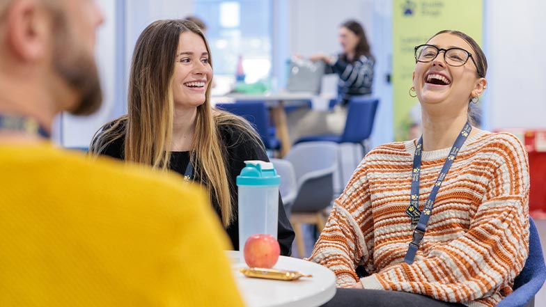 Three Guide Dogs employees sitting at a round table in a casual, open workspace, laughing and smiling