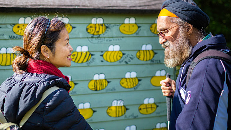 Pardeep who has sight loss is with his my guide volunteer Sophia smiling next to a shed painted with bees
