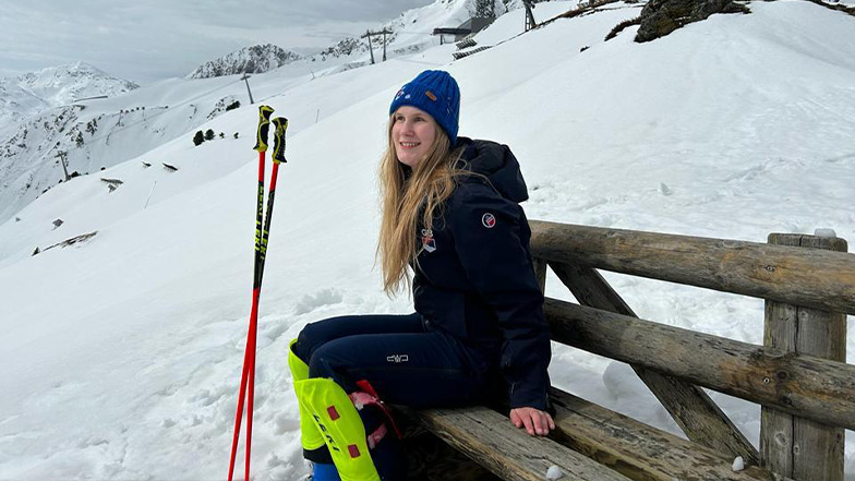 Hester sits on a bench smiling, on a snow covered ski slope.