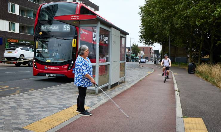 A photo of a Bus stop bypass where a long cane user is standing at the tactile paving on the edge of the bypass island waiting to cross over the cycle lane. There is a cyclist approaching about 4 metres away.