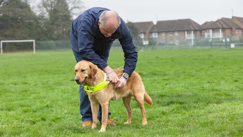 A guide dog owner and his guide dog stand in an open park, as he removes his dog's guiding harness.