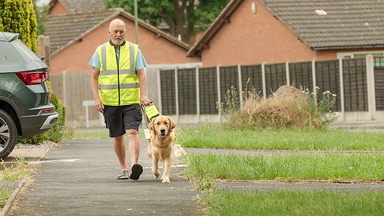 A guide dog owner walks along a pavement with his guide dog by his side.