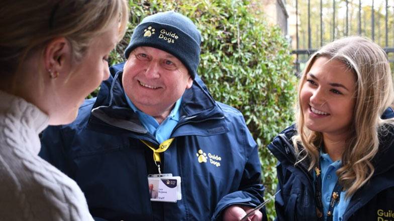 2 Guide Dogs' Fundraisers wearing ID badges speaking to a woman 