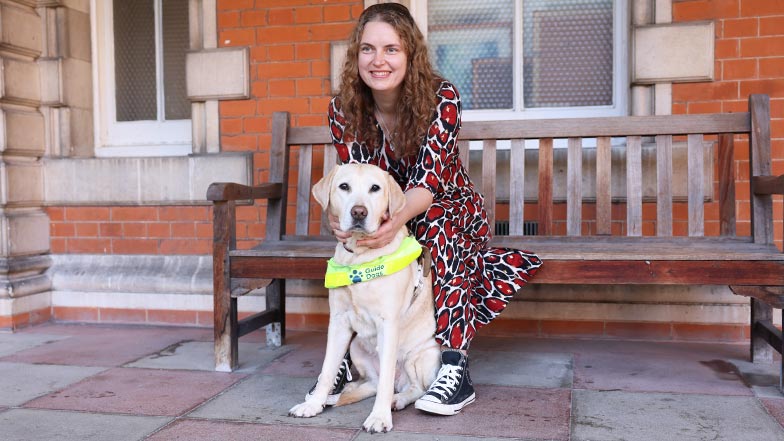 Smiling guide dog owner sitting on a bench with Labrador guide dog in harness at her feet