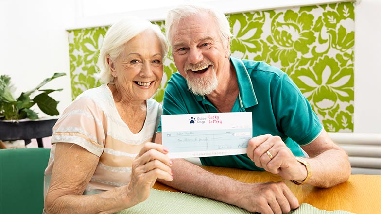 A smiling man and woman sitting at a table holding a Guide Dogs' Lucky Lottery winners cheque.