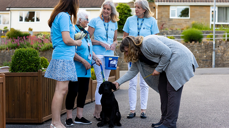 Guide dogs volunteers stand with a guide dog and member of public, smiling