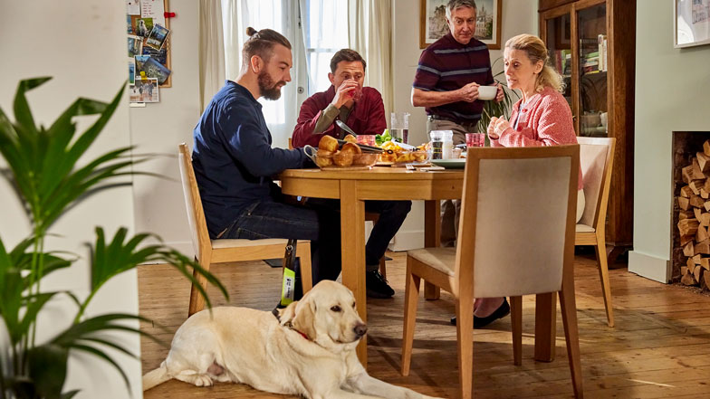 A family of four sit around a dining table with a guide dog laying at their feet.