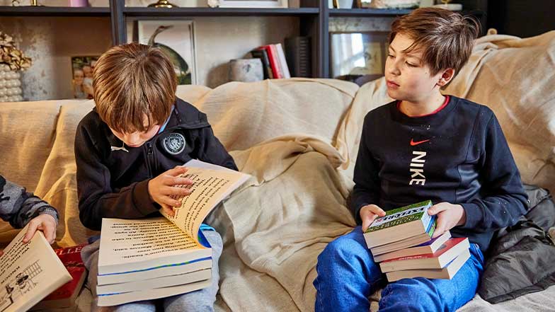 Two brothers sit on the sofa holding books. One of the brothers has a book with enlarged text.