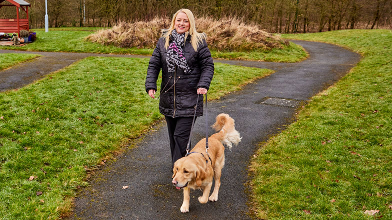 Guide dog Ringo and volunteer Louise walk along a path in a park.
