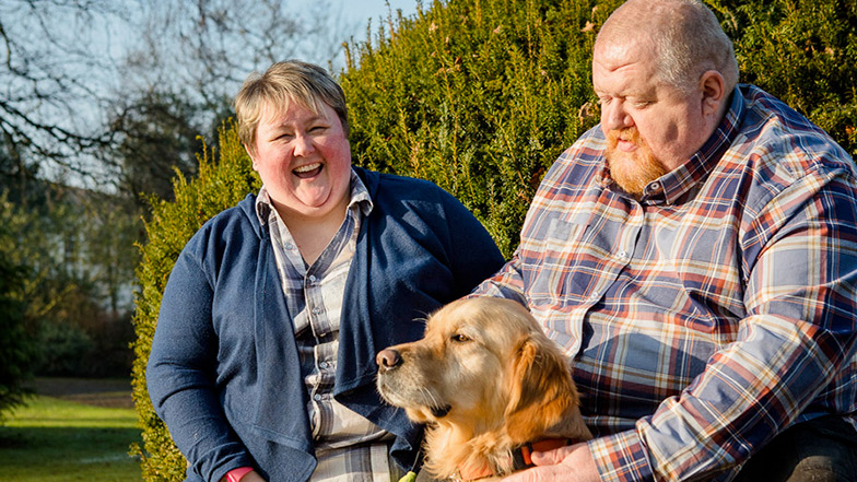 Two people sitting on a park bench with guide dog and smiling