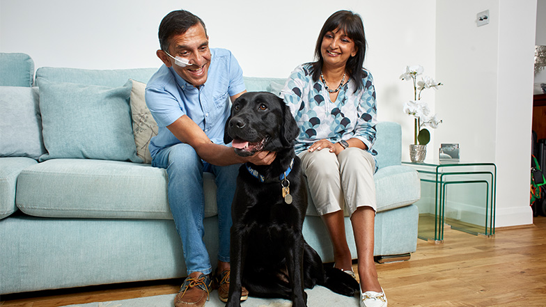 Two volunteer Fosterers sit on a sofa smiling whilst one strokes a black labrador dog