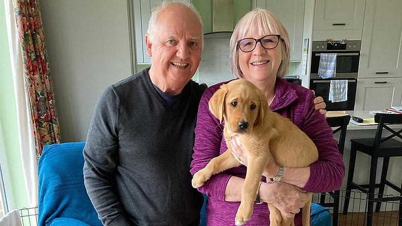 Volunteer puppy raisers hold guide dog puppy Titch whilst smiling