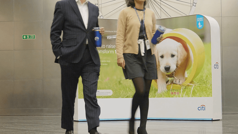 Two Citi employees walking through the Citi lobby with a Guide Dogs poster in the background
