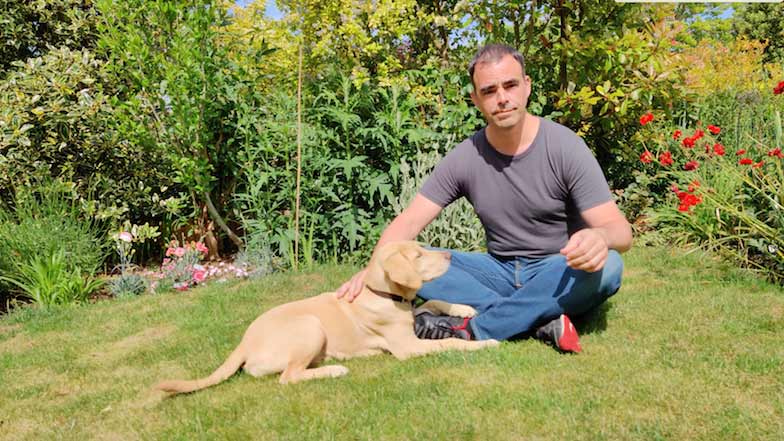 William, the guide dog puppy raiser sits with guide dog puppy Theia on the grass in their garden