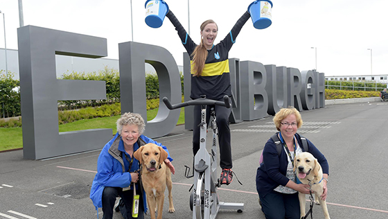 Two guide dogs in training and their handlers beside a girl sat on an indoor cycle, fundraising at Edinburgh airport