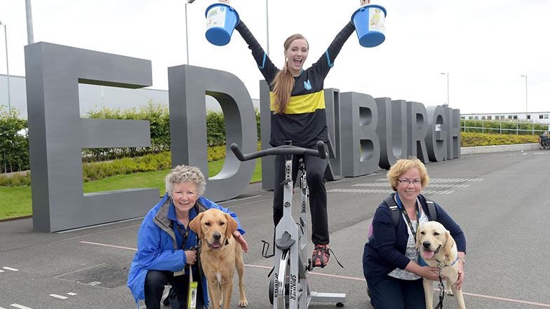 Edinburgh Airport staff member sits on an indoor cycle with two collection boxes and two guide dogs in training beside her