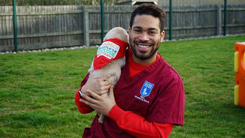 Danyl Johnson looks to camera. He is holding a guide dog puppy who is wearing a People's Postcode Lottery bandana
