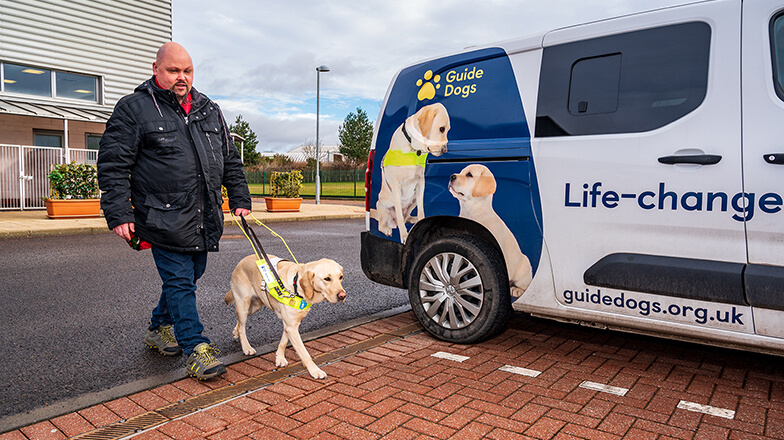 Guide dog owner Fraser walking past a Guide Dogs van with guide dog Mabel