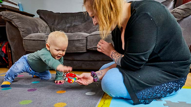 Young child Alfie is playing with a multi-coloured tambourine on the living room floor with his mum