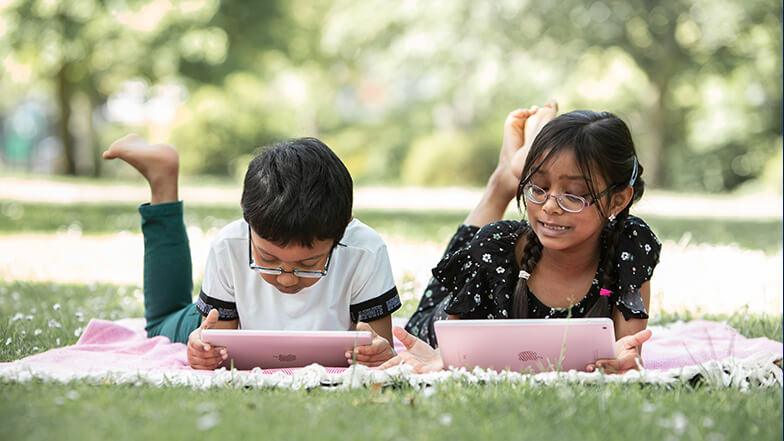Two children laying on the grass using ipads