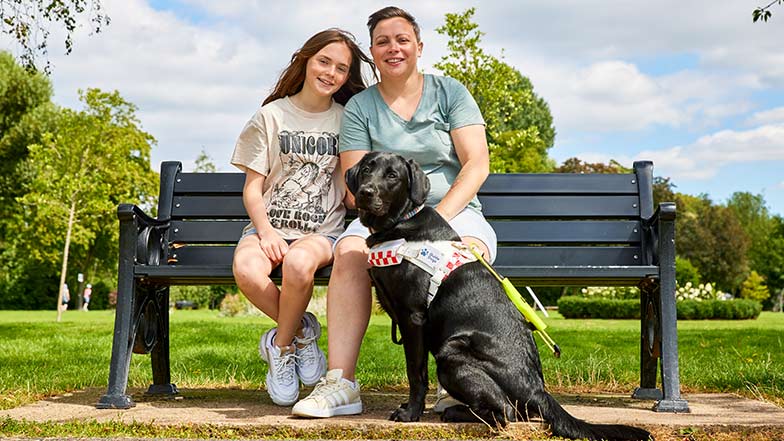 Claire and her daughter sitting on a bench smiling with guide dog Jacqui sitting in front of them