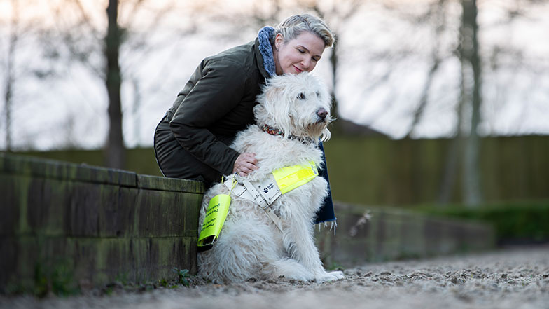 Guide dog owner Verity sitting with her hand lightly touching guide dog Luna outside.