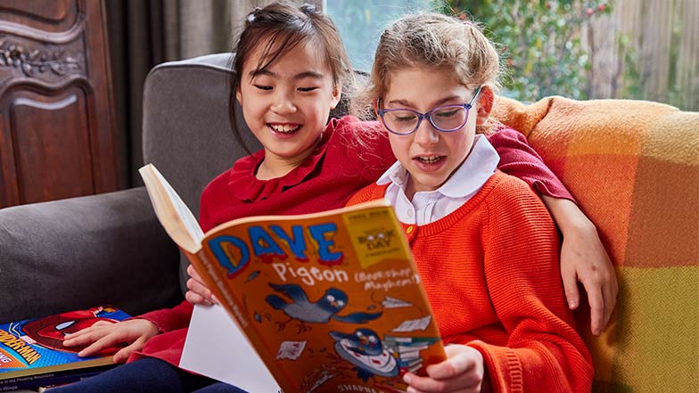 2 girls sitting huddled on a sofa enjoying a large print book.