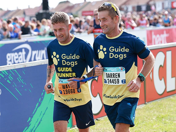 Darren Swales running the Great North Run with his brother as his guide runner, both wearing Guide Dogs vests