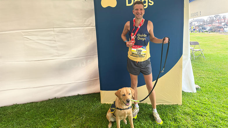 A man at a sporting event wearing a Guide Dogs running vest and standing with a Labrador