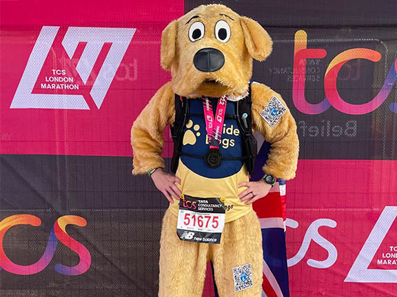 Matt is wearing a Guide Dogs t-shirt and dog mascot suit in front of London Marathon banner