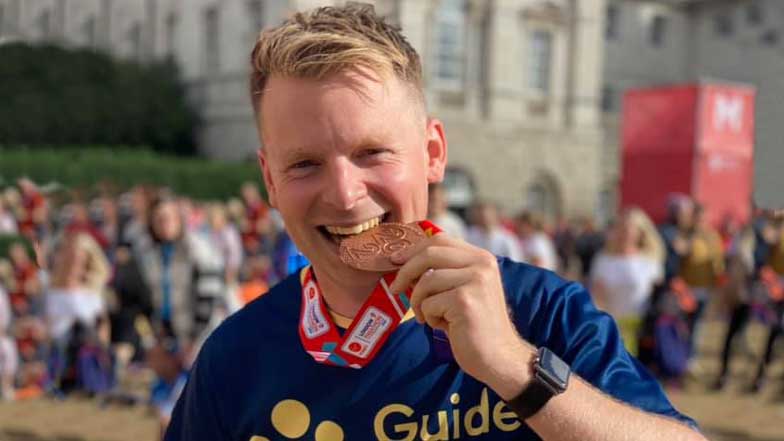 A man smiling and biting down on his medal after running a challenge event for Guide Dogs