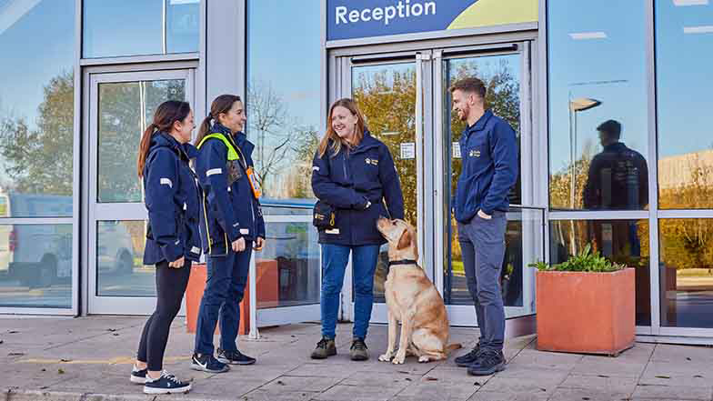 Four Guide Dogs staff members at a centre, with a Labrador sitting beside one of them.