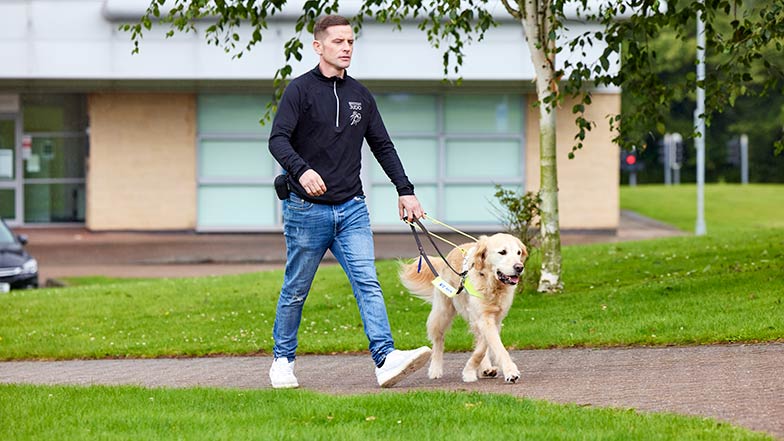 Guide Dog Owner Scott walks in a park with his guide dog Milo.