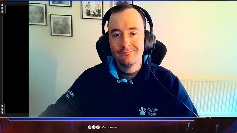 Screenshot of Scott streaming on his Twitch channel as Velcromad. He is smiling at the camera and wearing headphones and a dark blue Guide Dogs hoodie.