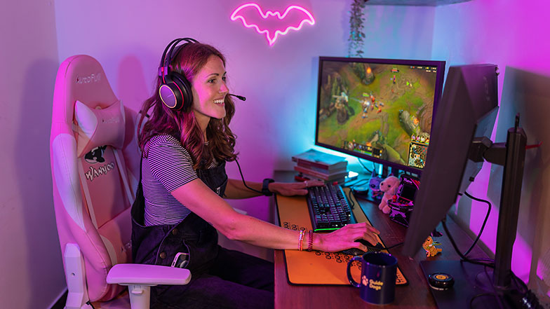 A smiling woman wearing a headset sitting in front of a gaming PC. The room is lit in bright pink and blue neon lighting