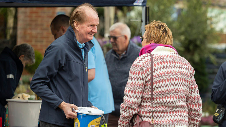 A Guide Dogs volunteer with a collection bucket talking to someone