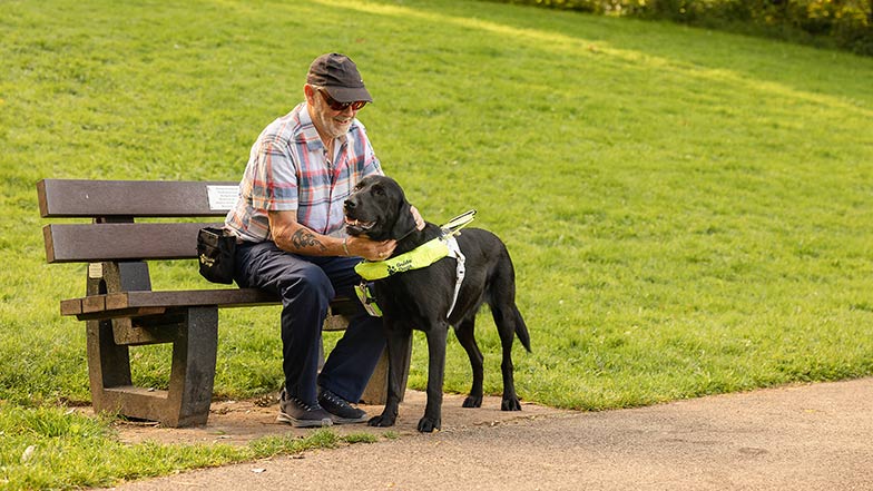 A guide dog owner sitting on a bench smiling with their guide dog by their side