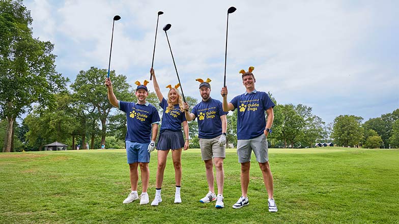 Four people on a golf course waving clubs in the air and wearing Guide Dogs T-shirts and dog ears