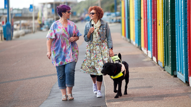 Two ladies walking down a beach promenade with their guide dog