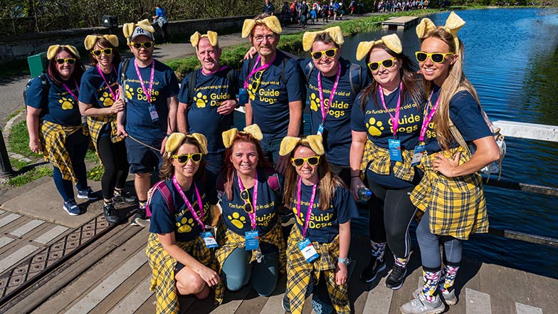 A group of Guide Dogs Kiltwalker’s wearing dog ears and Guide Dogs t-shirts smile for a photo on a bridge.