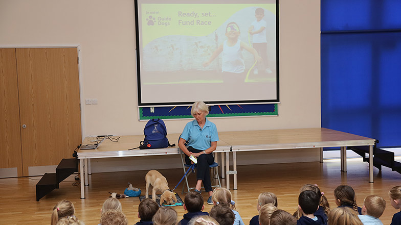 A Guide Dogs speaker with their dog at a school assembly with a Fund Race presentation behind them