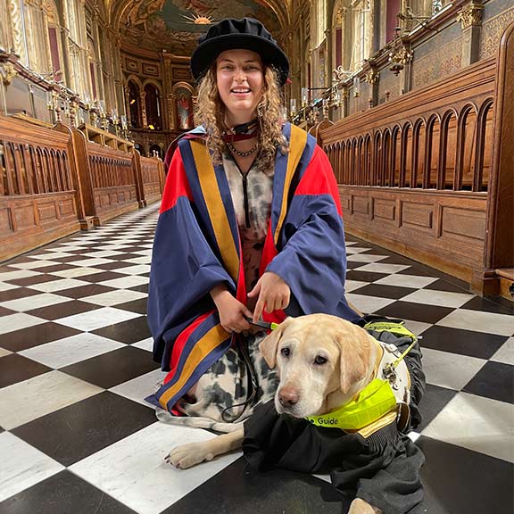 A smiling Anica in her graduation robes with guide dog Lassie at her feet