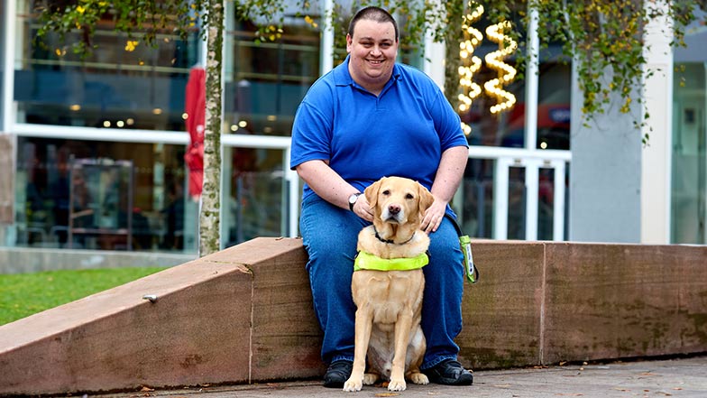 Smiling guide dog owner Charlies sitting on a wall with guide dog Carlos in harness sitting at his feet.