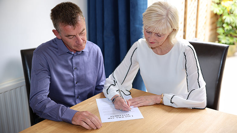 A man and a woman sit at a table looking at an A4 piece of paper, the woman has a pen in her hand.