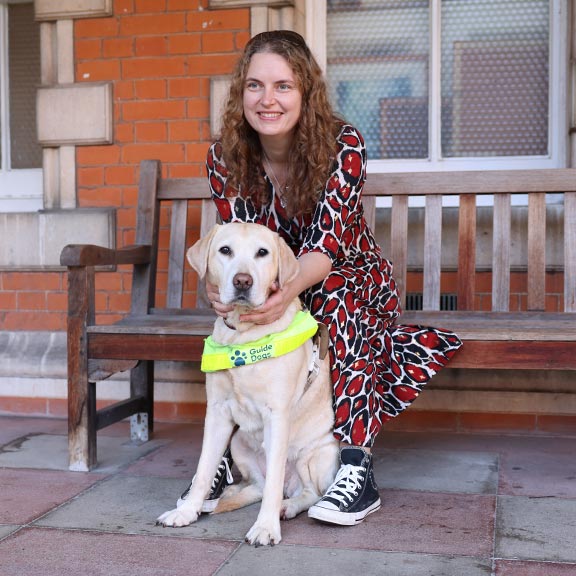 Guide dog owner Anica sitting on a wooden bench with guide dog Lassie sitting in front in harness
