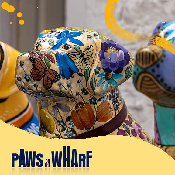 Close up of the head of a decorated guide dog sculpture. It is painted gold with multi-coloured flowers and wildlife. There is a yellow banner with the Paws on the Wharf logo.