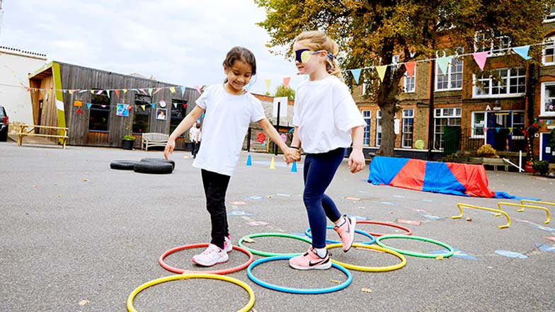 Two girls on the school playground, one is blindfolded and the other is helping her through some hoops on the ground