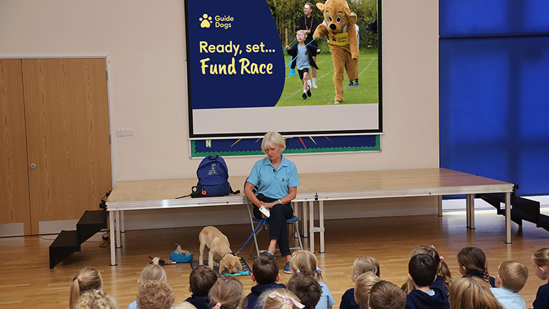A Guide Dogs speaker talking in a school assembly with a presentation on Fund Race.