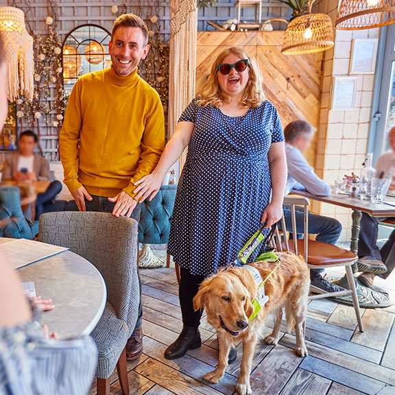 A man who is a sighted guide, guides a woman with vision impairment to a table in a bar, her guide dog is also there.
