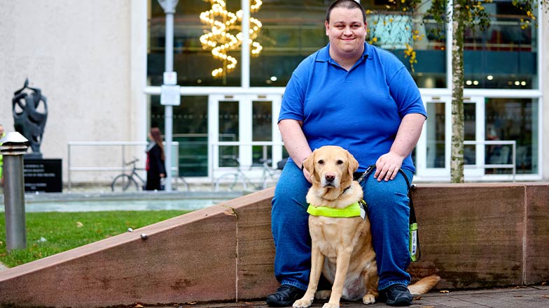 Guide dog owner, Charles, sits on a low wall with his guide dog at his feet.
